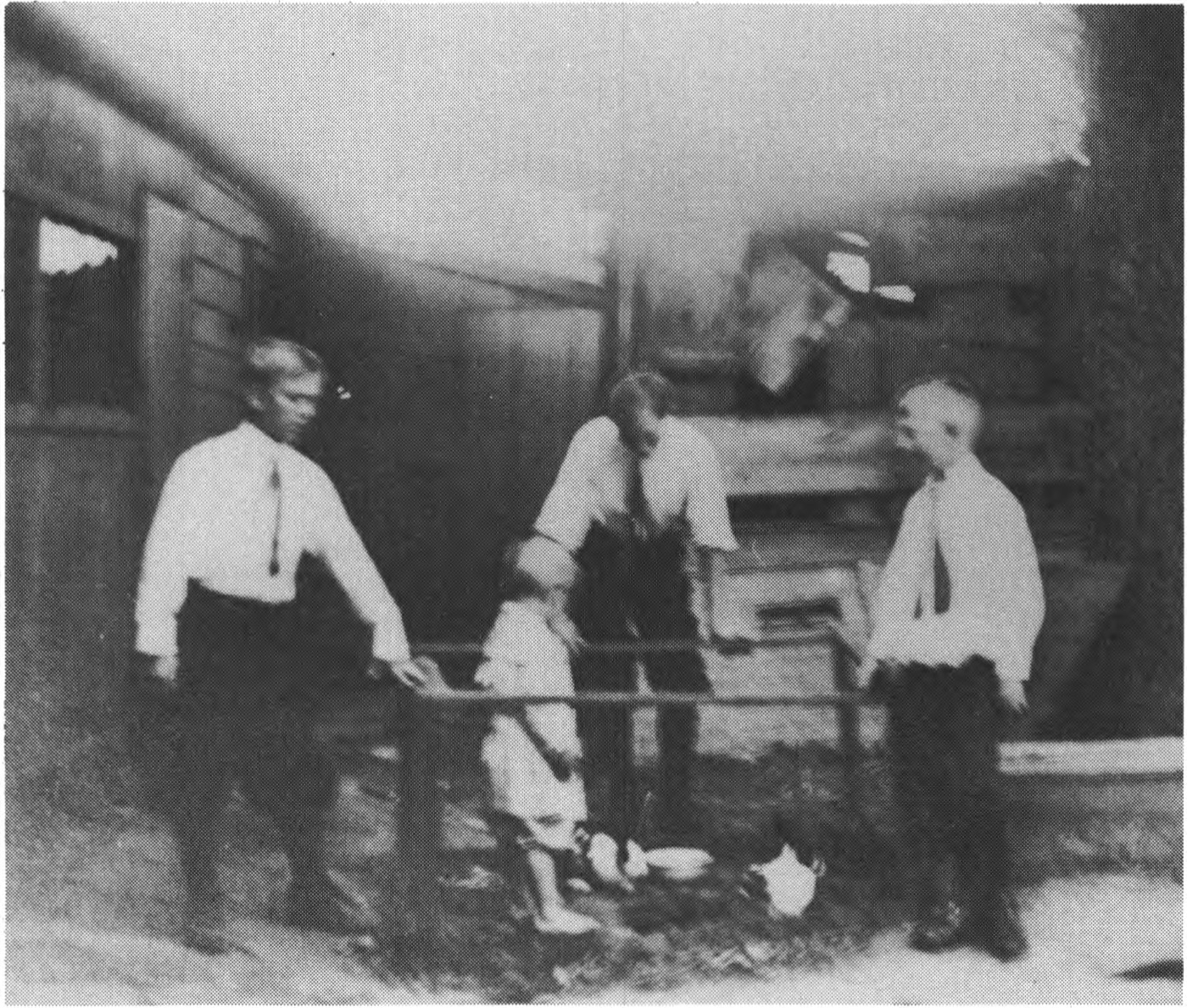 Four boys in white shirts and neckties in the yard behind a clapboard house; three stand along a pipe rail facing the camera and a fourth, in a cap, looks over from behind them.