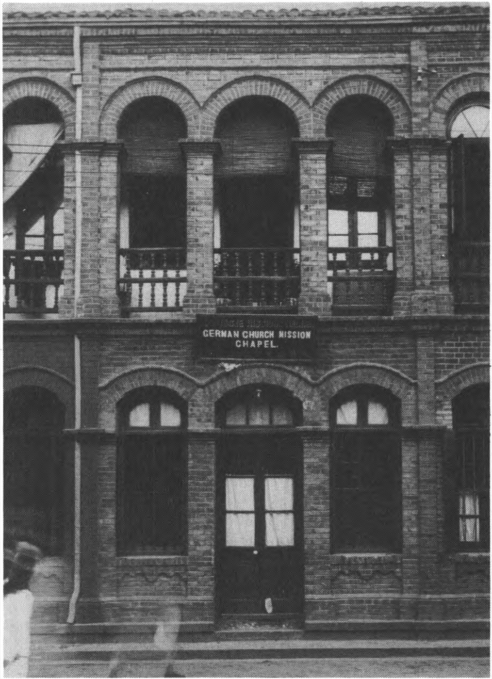 A two-storey brick building with round-arched windows and a balustraded balcony; a painted board fixed between the floors reads GERMAN CHURCH MISSION CHAPEL.