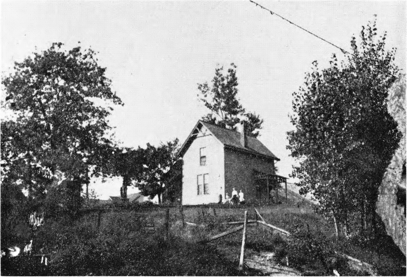 A plain two-storey frame house standing on a rise, photographed from across a rough yard with a broken post-and-wire fence and trees on either side; two small figures sit near the porch.