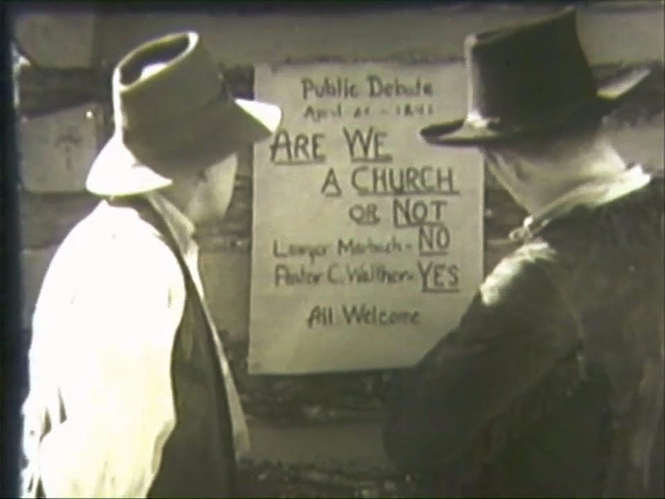 Two men in hats, seen from behind, read a hand-lettered placard on a log wall: Public Debate, April 21, 1841. Are we a church or not. Lawyer Marbach, No. Pastor C. Walther, Yes. All welcome.